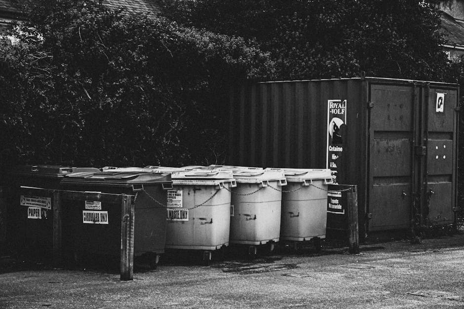A row of large wheeled rubbish bins lined up along a pavement adjacent to a brick wall, with green, blue, red, and dark green coloured lids and bodies, each with a textured lid surface. The bins are p
